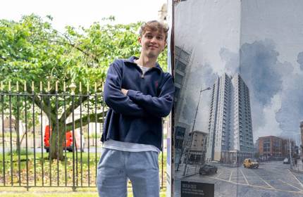 A young man crosses his arms and leans against a large poster displaying Cardiff architecture