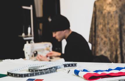 A close-up of a measuring tape and piece of striped fabric. In the background, a person operates a sewing machine.