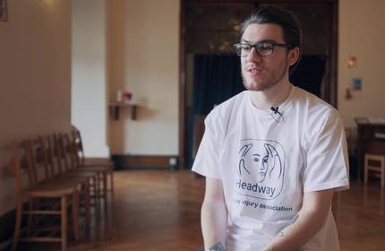 Sam Wilson, wearing a white t-shirt and glasses, sits in a reception room