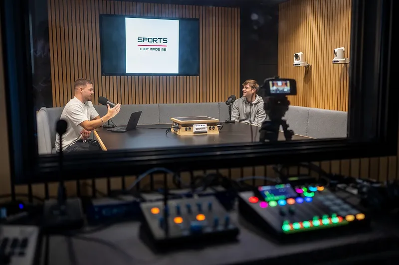 Two young adults sit at far ends of a table talking to one another recording a podcast in a sound booth.