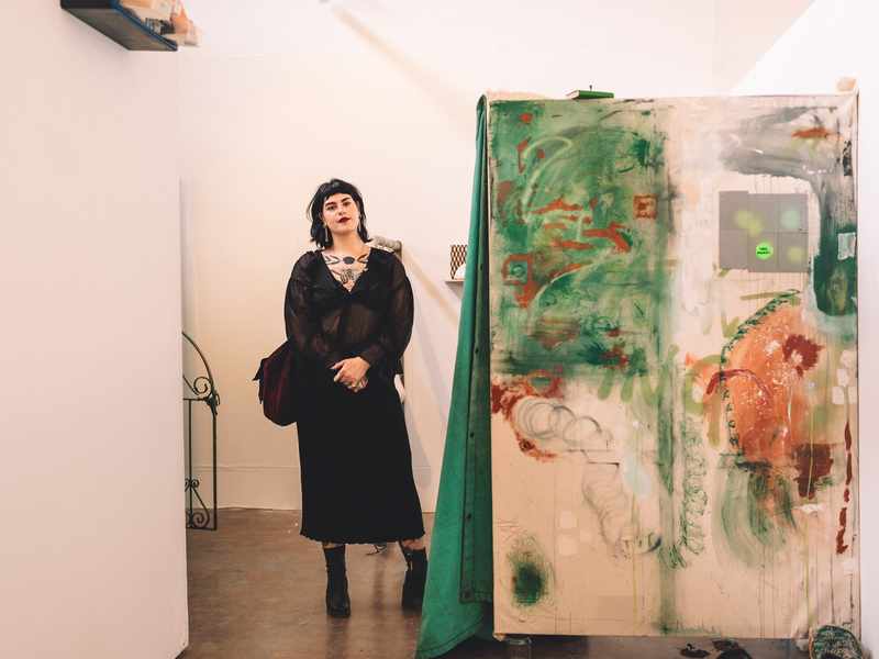 A young woman in a black dress stands beside a large painted canvas