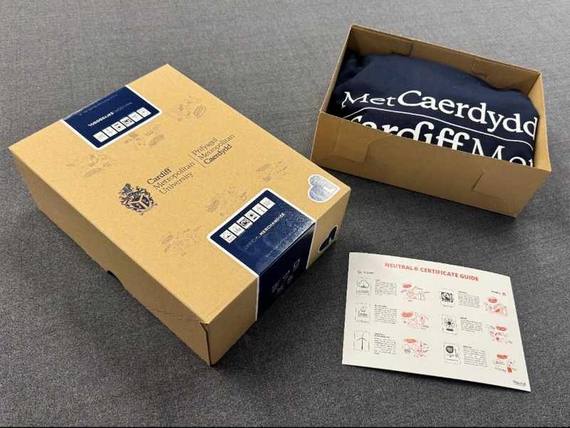 Two cardboards boxes, one opened and containing a Cardiff Met navy hoodie, on a carpeted floor
