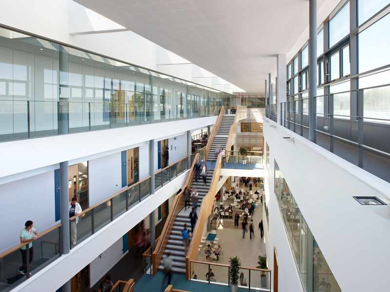 A modern, spacious atrium with a large wooden staircase and multiple people walking. The area has glass railings, plants, and abundant natural light from skylights above. The scene conveys a busy, contemporary office or educational environment.