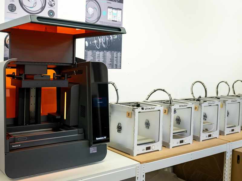 A collection of 3D Printers lined up on a row of desks