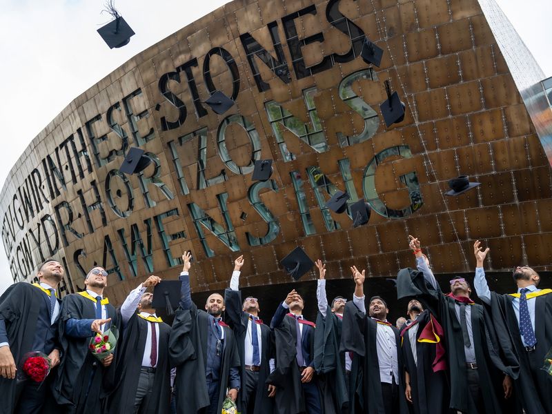 A row of student toss their graduation caps into the air in front of the Wales Millennium Centre sign and entrance