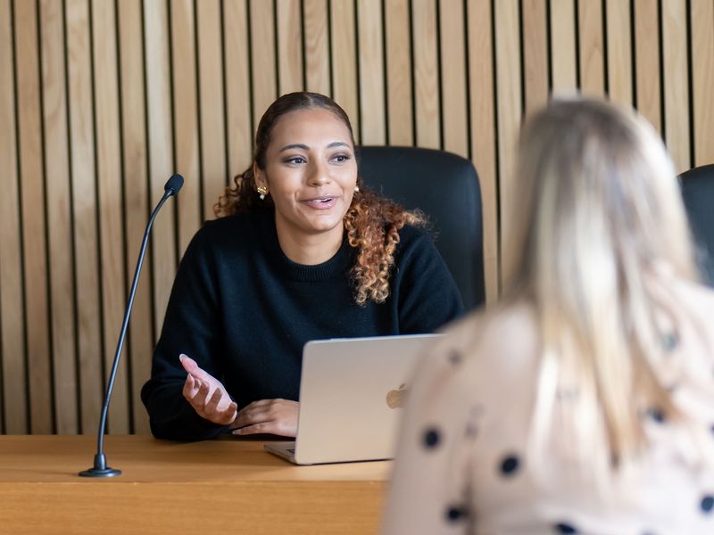 A person sits at a courtroom bench. They are in conversation with someone sat across from them.