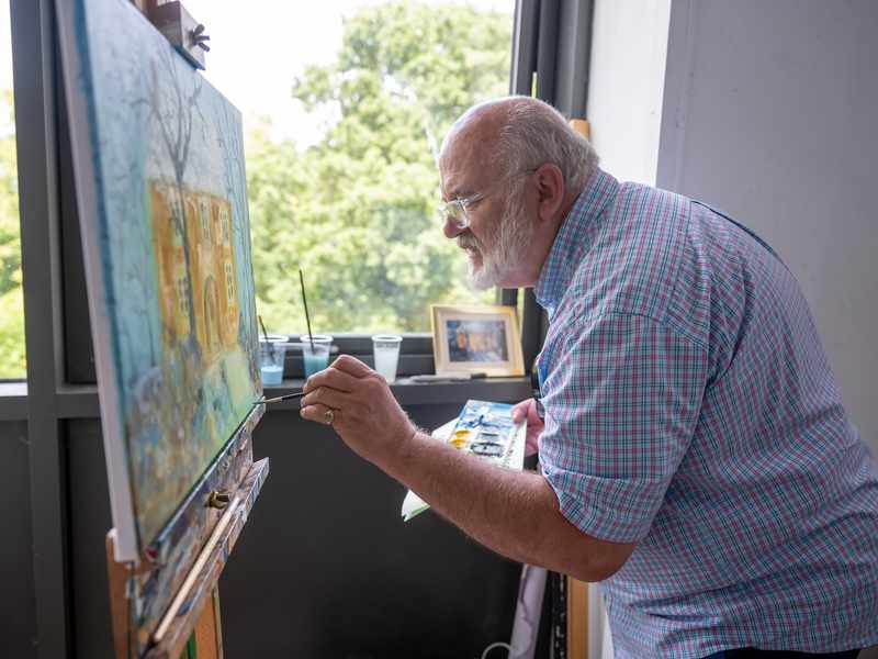 An elderly man in a short sleeve shirt paints on a large canvas