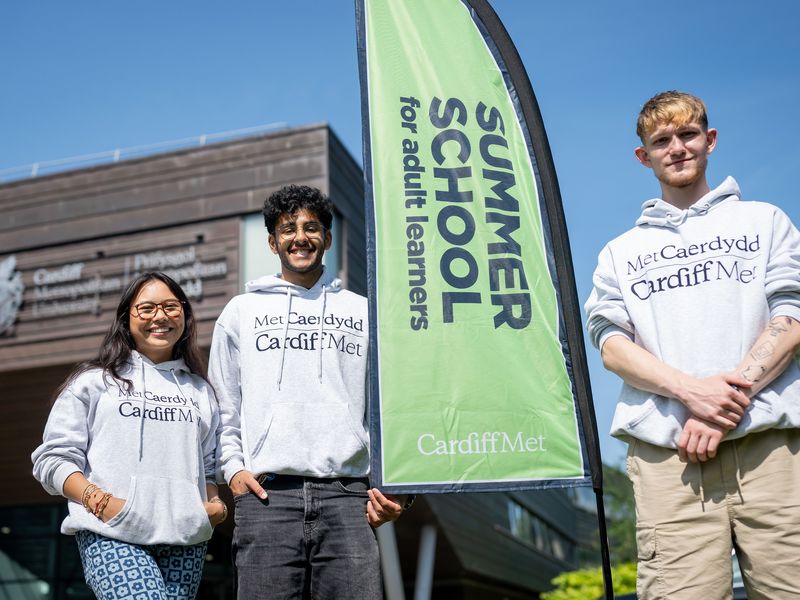 Three young adults in Cardiff Met hoodies stand beside a Cardiff Met Summer School banner