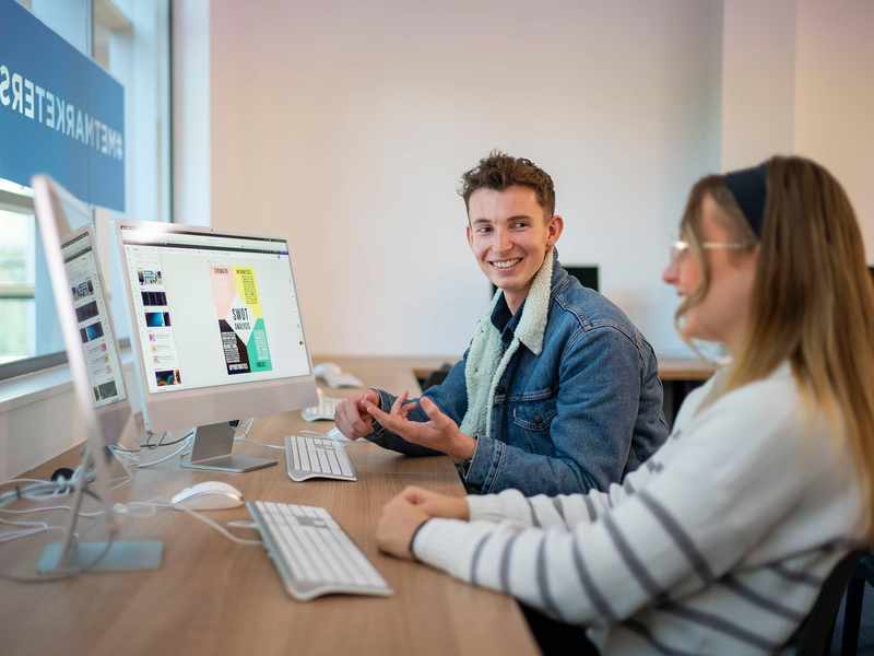A desk with two people working on computers. One person is talking to the other.
