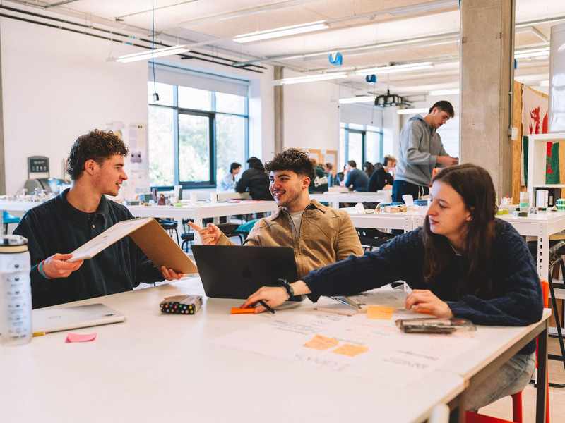 Three students sit together at a table in a studio space. One is holding a sketchbook, and there are laptops, pens and paper on the table.
