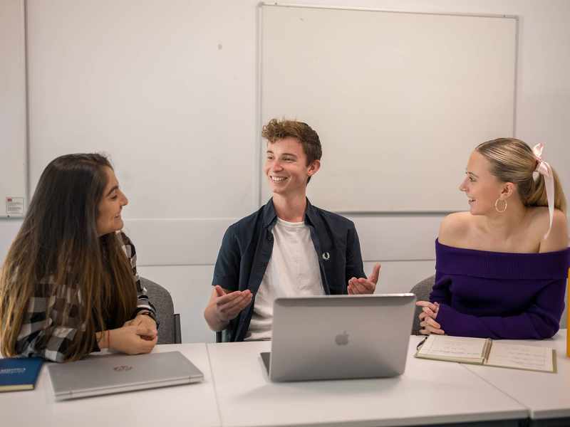 Three students sit alongside each other at a table with laptop computers and notebooks.