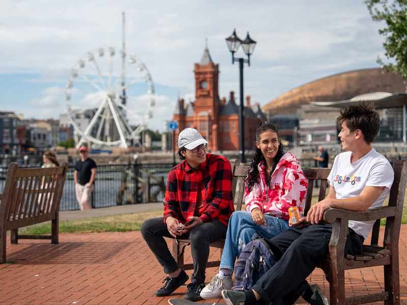 Three students sit on a bench in Cardiff Bay. Behind them is a Ferris wheel, the Pierhead Building and Wales Millennium Centre.