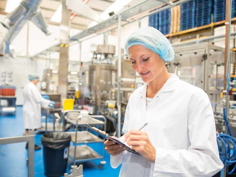 A woman in a white lab coat and hairnet writes on a clipboard in a factory