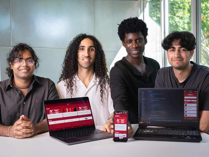 Four men smile for a photo. In front of them are two laptops showing an app in development. One is holding a mobile phone showing the same app.