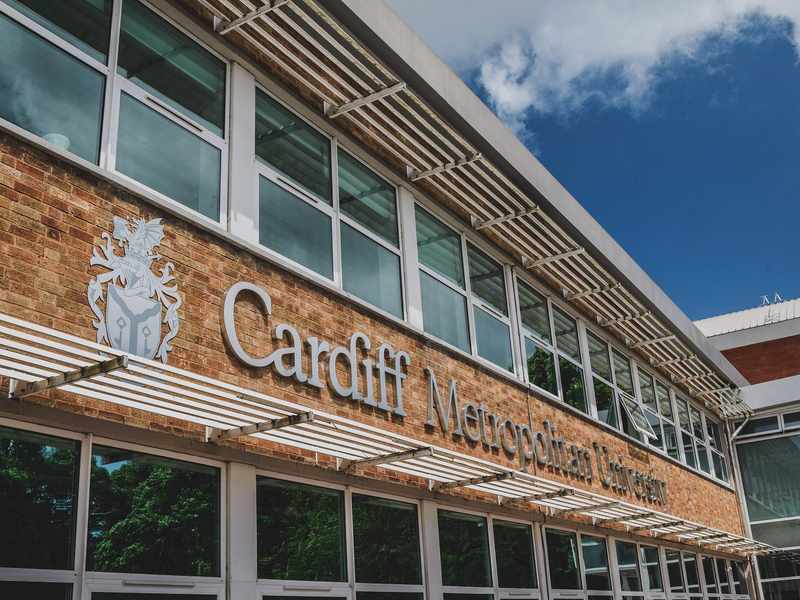 A building with large windows featuring the sign Cardiff Metropolitan University on a brick wall. A blue sky with clouds is visible above.