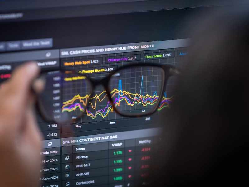 A computer screen displaying stock market data and graphs, viewed through a pair of glasses held in someone's hand.