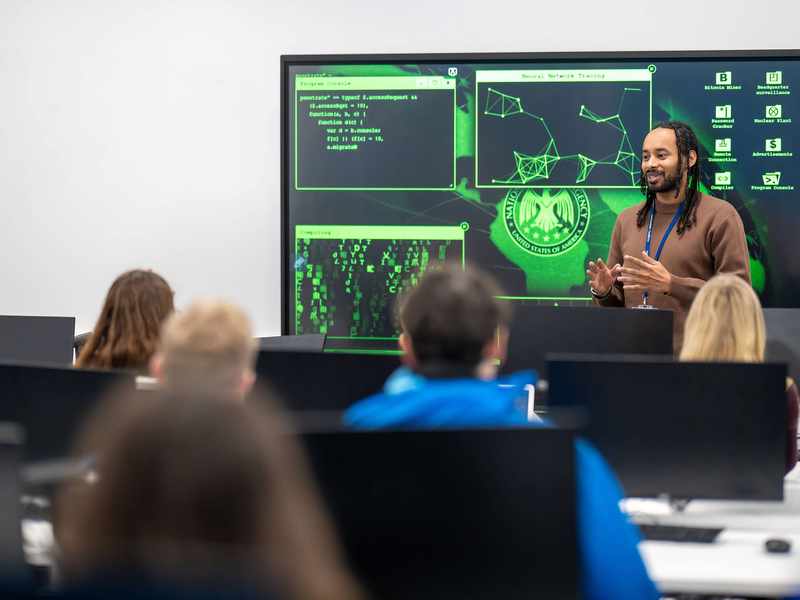 A lecturer stands in front of a large screen displaying computer graphics, engaged in a presentation or discussion.
