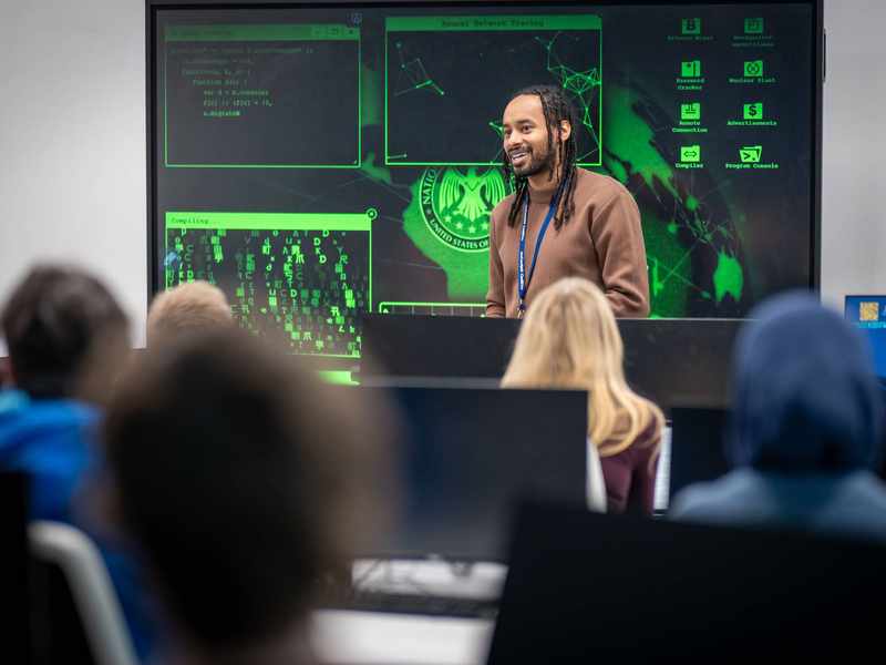 A lecturer stands in front of a large screen displaying computer graphics, engaged in a presentation or discussion.