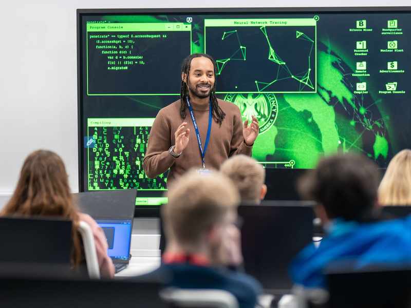 A lecturer stands in front of a large screen displaying computer graphics, engaged in a presentation or discussion.