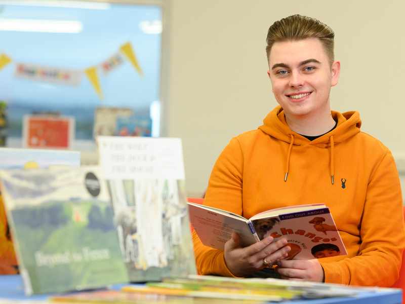 A person in an orange hoodie is smiling while holding a book in a library. There are several books displayed in the foreground. The background shows blurred decor and furniture.