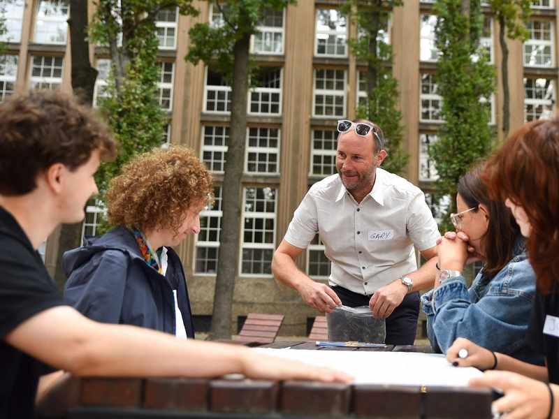 Four people sit at a wooden table outdoors. A man wearing a name badge labelled Gary stands next to them.