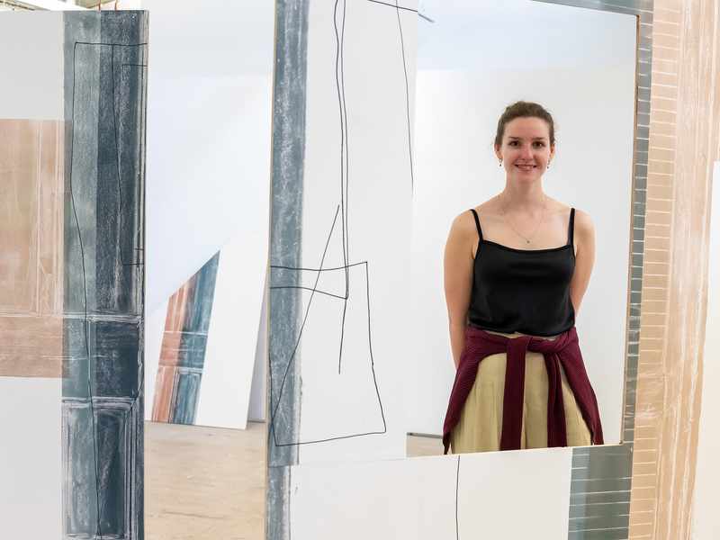 A woman stands smiling amidst an abstract art installation with geometric designs on large panels. She is wearing a black top and has a sweater tied around her waist. The setting appears to be an art gallery.