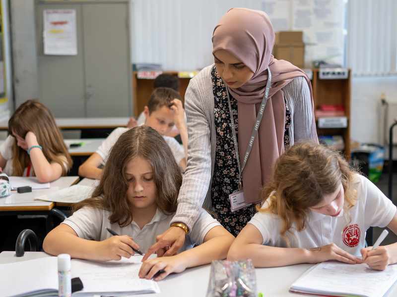 A teacher stands over two pupils writing at their desks.
