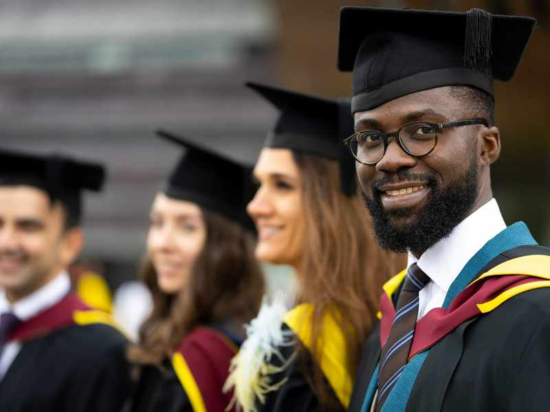 A graduate in cap and gown. In the background are three other graduates also wearing caps and gowns.