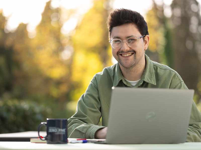 A young man sitting outside at a table. In front of him is a laptop computer and coffee mug.