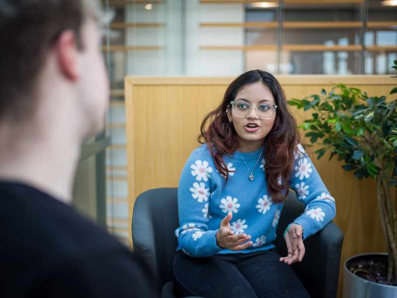 A young woman with glasses and long hair, wearing a blue sweater with white flower patterns, gestures while talking. She is seated in a modern indoor space, facing another person whose back is to the camera. A green plant is in the background.