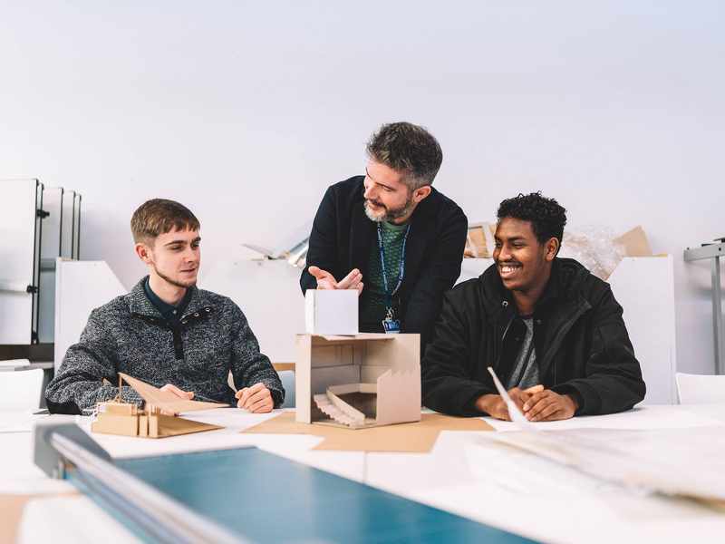 A lecturer stands between two students seated at a table with small architectural models made from card.