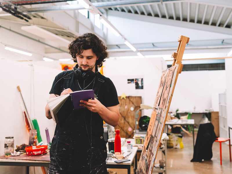 A young man standing next to an easel writes in a notebook.