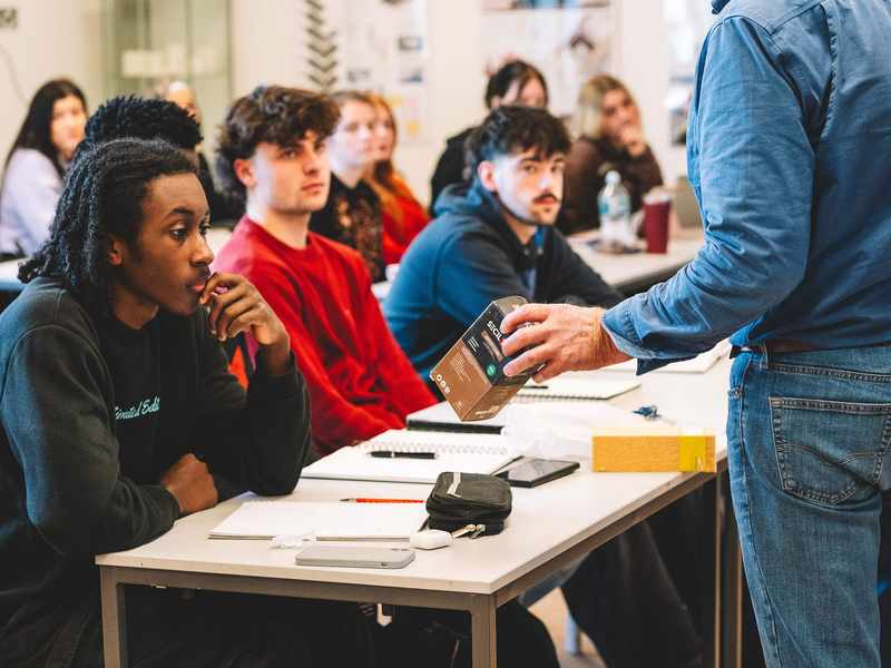 Student being shown a boxed product while seated at a classroom table with others listening nearby.