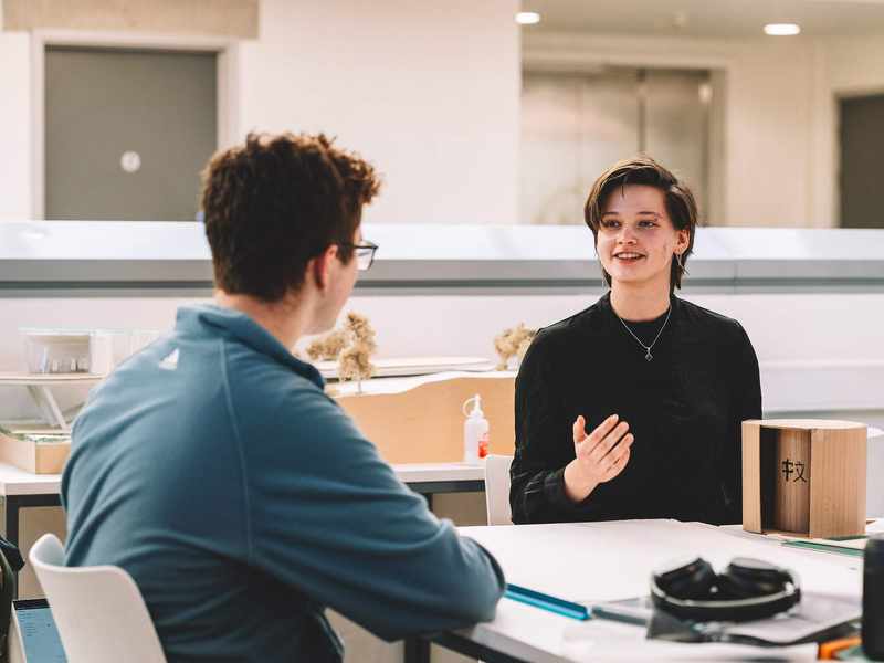 Two students seated at a table discussing a small cardboard model with tools and materials nearby.