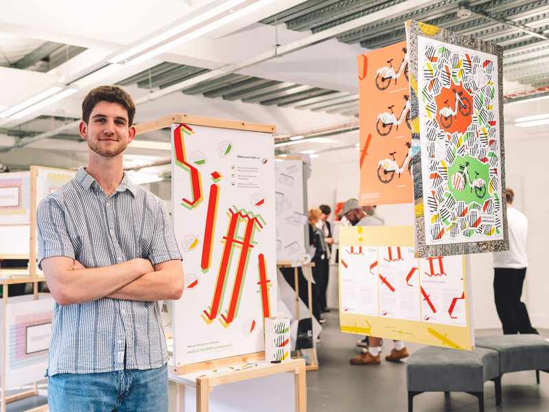 A student stands with arms folded next to a display of design work.
