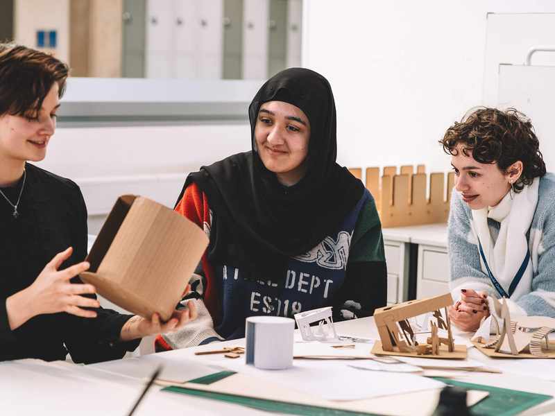 Three students seated at a table reviewing small architectural models and cardboard structures.