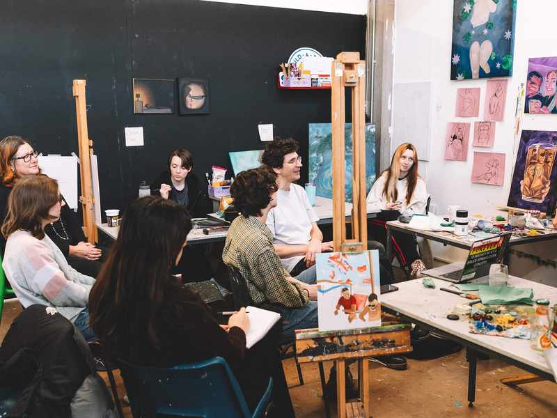 A group of students in a studio space, surrounded by canvas paintings and sketches.