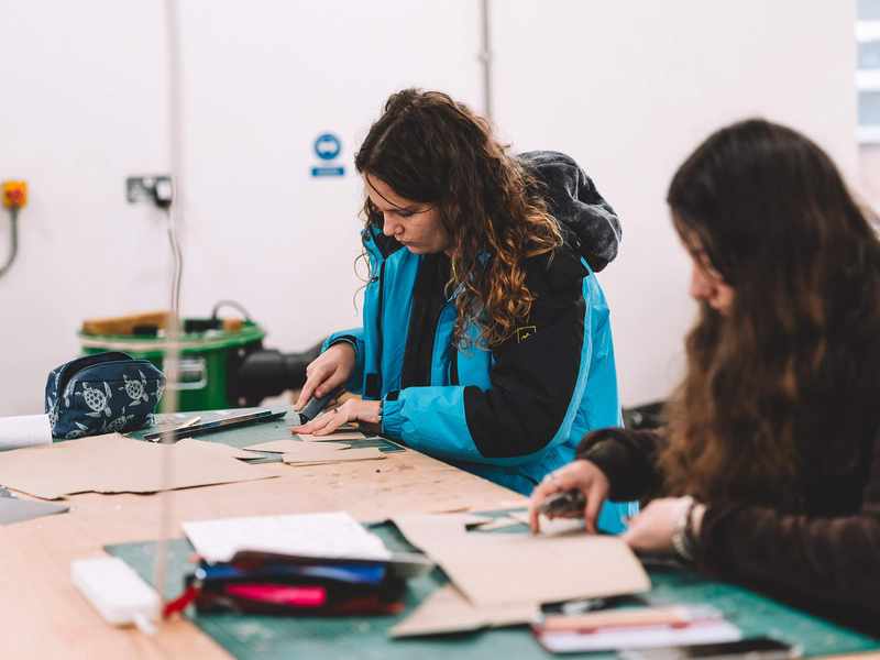Students cutting cardboard pieces on a workshop table with craft tools and materials spread out.