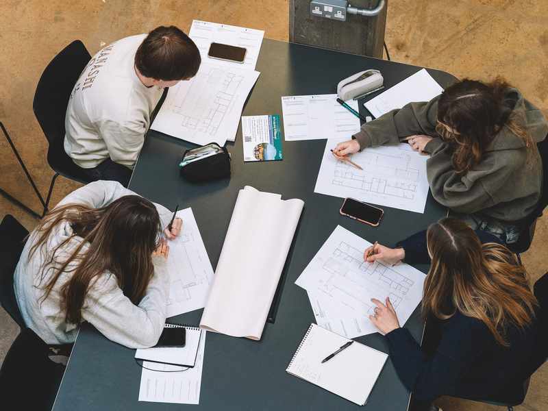 Group of students seated around a table studying architectural plans with notebooks and phones nearby.