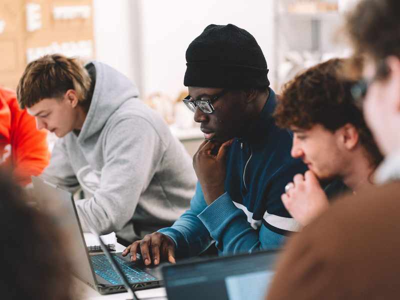 Students seated at a table working on open laptop computers in a studio environment.