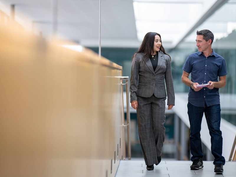 Two people walk alongside each other at the top of a staircase. In the background are glass walls overlooking an open atrium space.