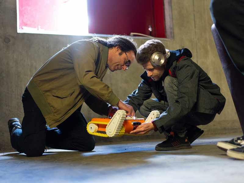 A person holds a small robot with caterpillar tracks while another person adjusts wires on its casing. They are in a room with a bare concrete floor and walls.