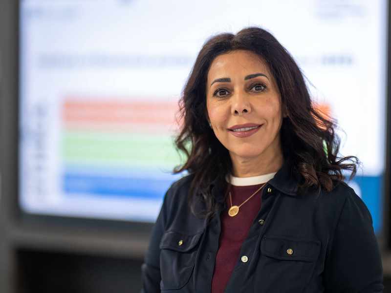 A woman wearing a dark blue jacket and maroon sweater. In the background is a large display screen.