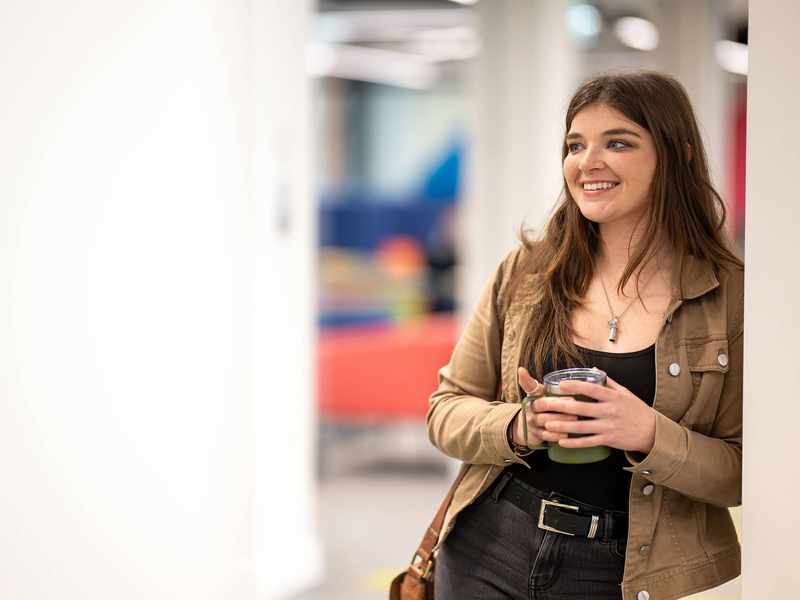 A young woman leaning against a wall, holding a reusable coffee mug.