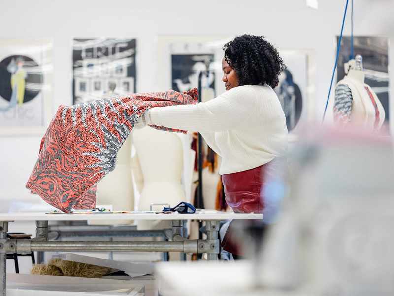 A person with curly hair holds up a colourful fabric in a design studio. In the background, a mannequin and framed art are visible. The room is well-lit, creating an inspiring workspace for creativity.