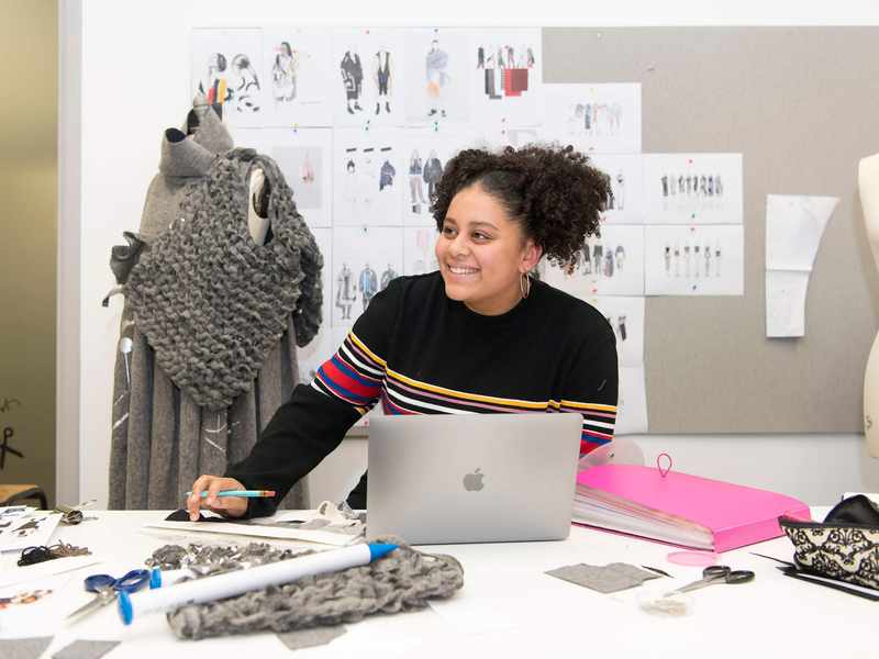 A young woman sits at a desk with a laptop computer and selection of fabric swatches. Behind her is a mannequin and wall of clothing designs.