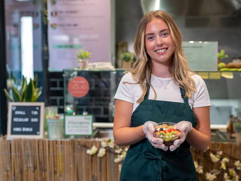 A young person wearing a green apron holds a fruit bowl while standing in front of a kitchen counter.