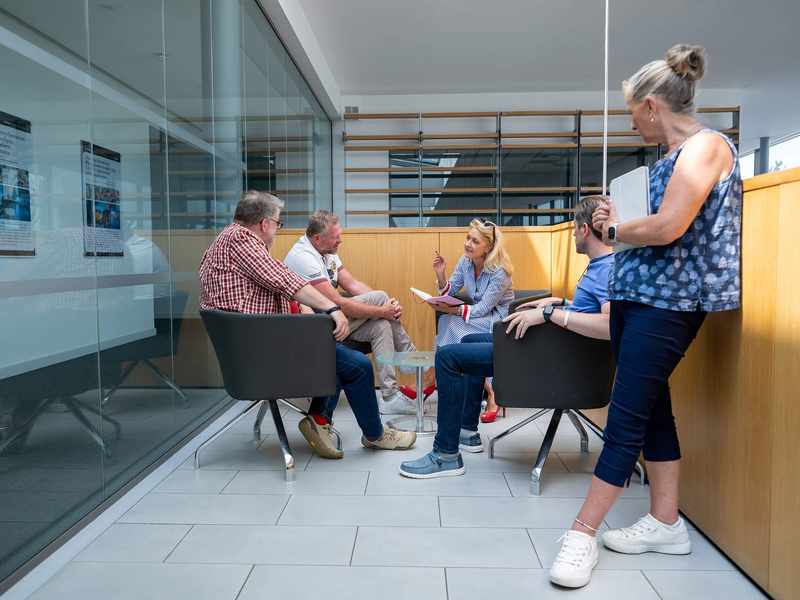 A group of people sit around a small table engaged in conversation. They are in a meeting space with a glass wall. A corridor is visible through the glass.