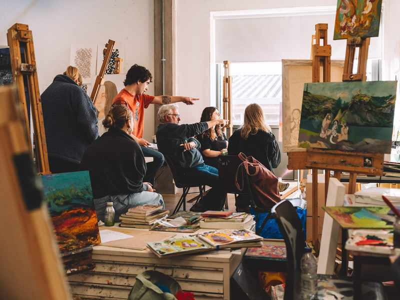 A group of students study a canvas painting mounted on an easel. There are other canvasses on the tables around them, along with sketchbooks.