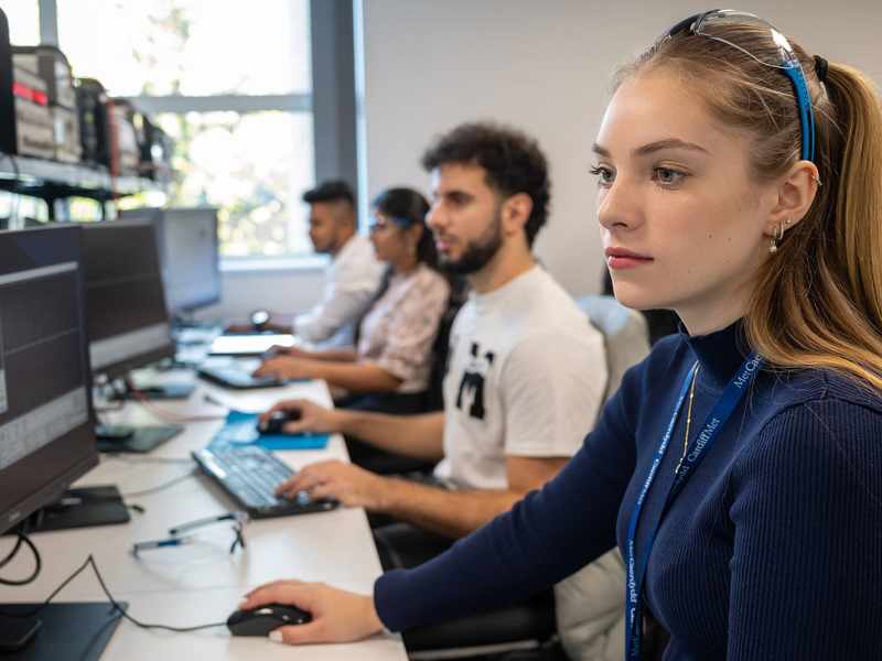 A group of people sit alongside each other at a long desk. Each of them is using a computer workstation.